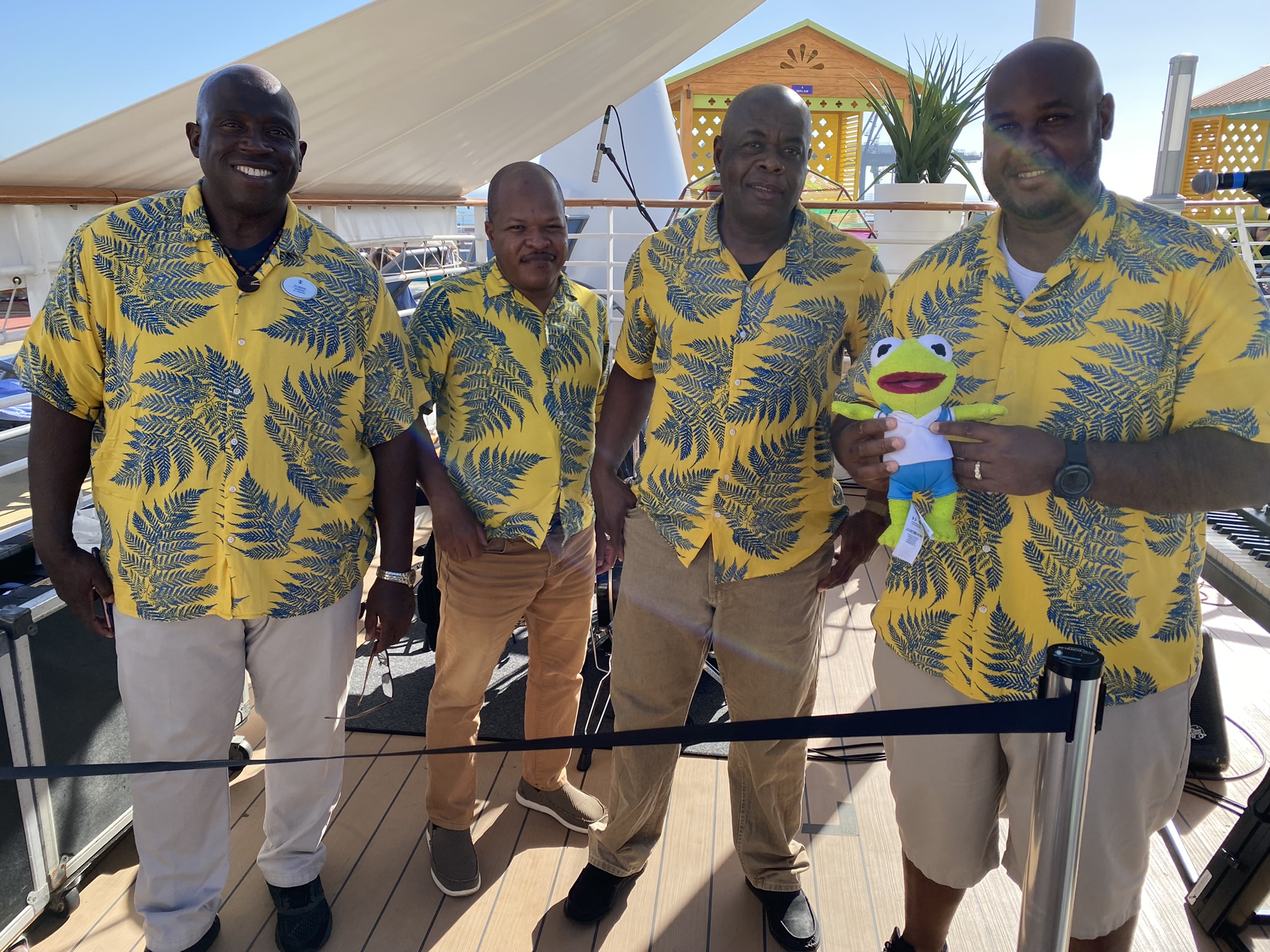 Kermit being held by one of the four musicians who are all wearing yellow hawaiian shirts with palm tree leaves all over.